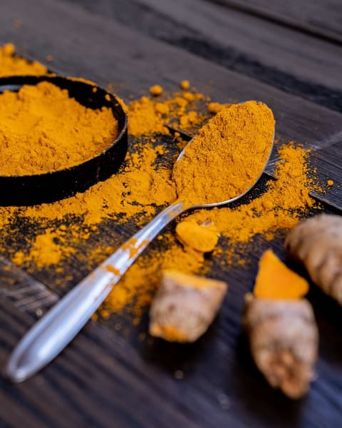 Fresh turmeric root and ground turmeric powder with a spoon on a dark wooden surface.