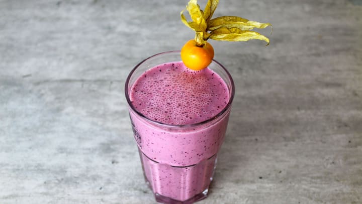 A creamy violet-pink blueberry milk smoothie in a faceted glass, its frothy top flecked with blueberry, on a moody stone surface.