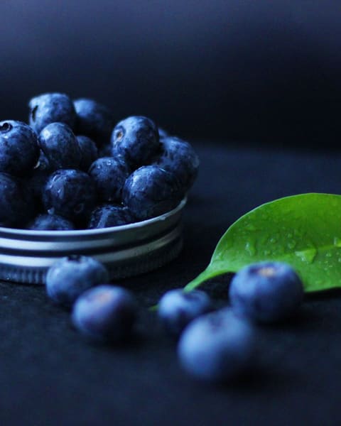 A small dish of fresh blueberries with a single green leaf on a deep navy background.