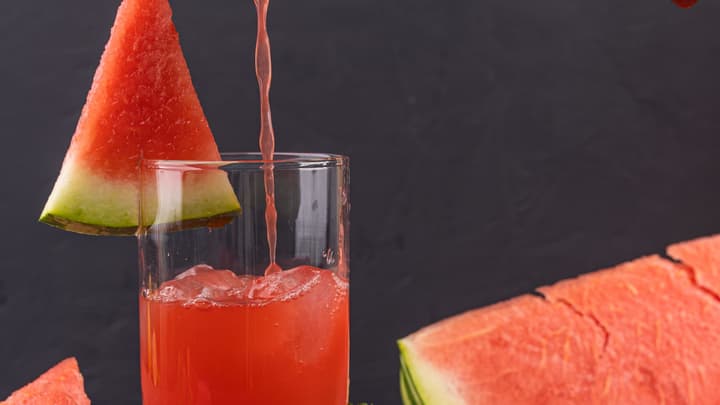 Bright pink watermelon juice pouring into a glass beside fresh watermelon wedges and mint, on a dark surface.