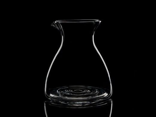 An elegant clear glass carafe with a delicate pour spout, glowing against a deep black background — a preview of the signature glassware collection.